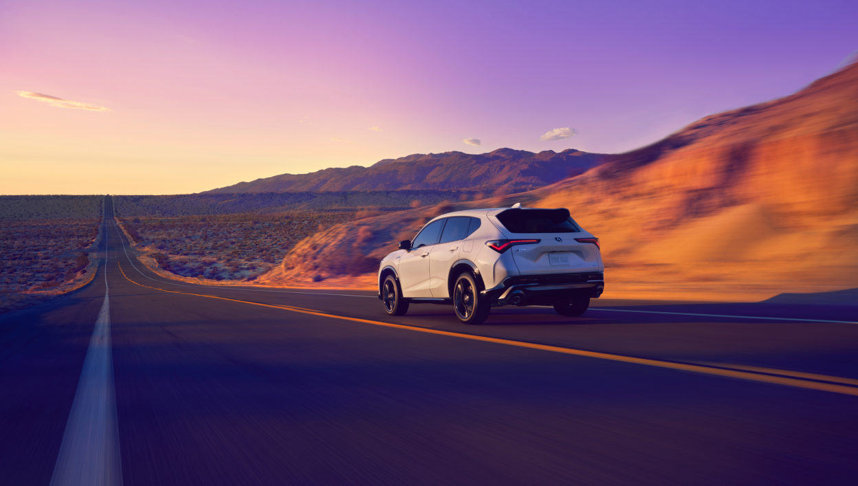 A back-view of a white 2025 Acura ADX driving on a long road surrounded by beautiful mountains and a purple-hue sky.