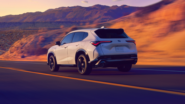 A back-view of a white 2025 Acura ADX driving on a long road surrounded by beautiful mountains and a purple-hue sky.