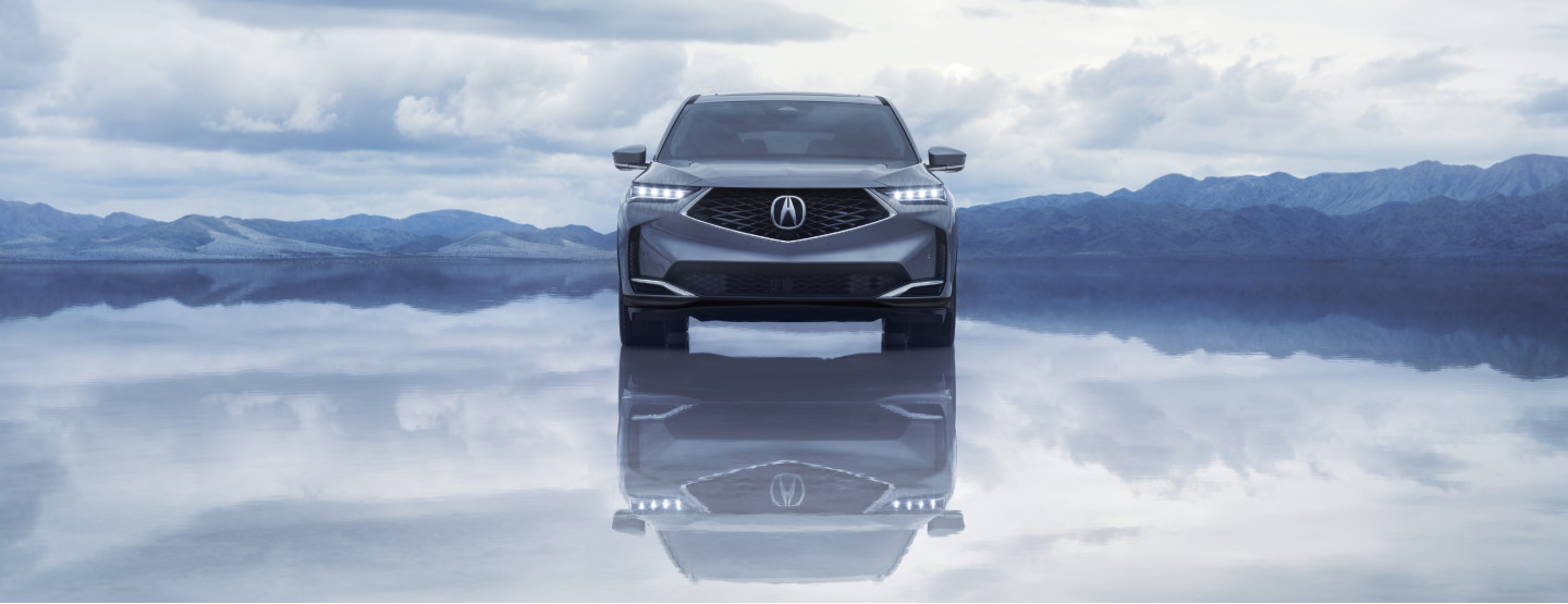 3/4 view of a grey MDX driving down a road in a mountainous desert landscape. / Vue 3/4 d’un MDX gris circulant sur une route dans un paysage montagneux désert.