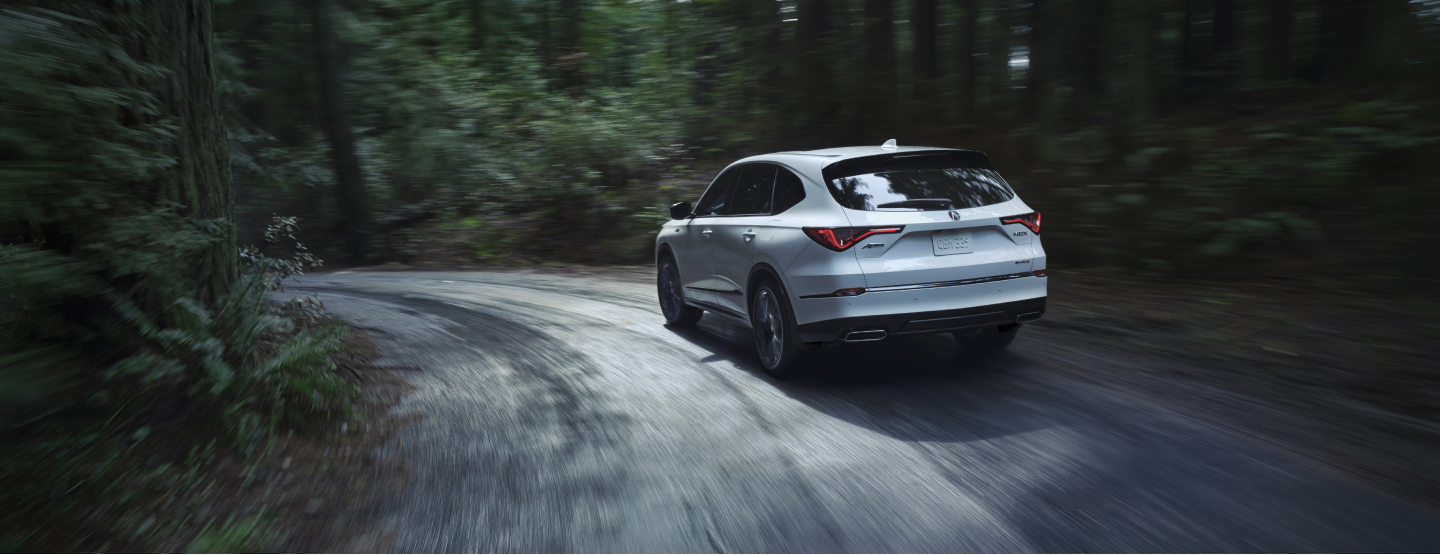3/4 rear view of white MDX taking a turn on a narrow concrete road in a temperate rainforest.