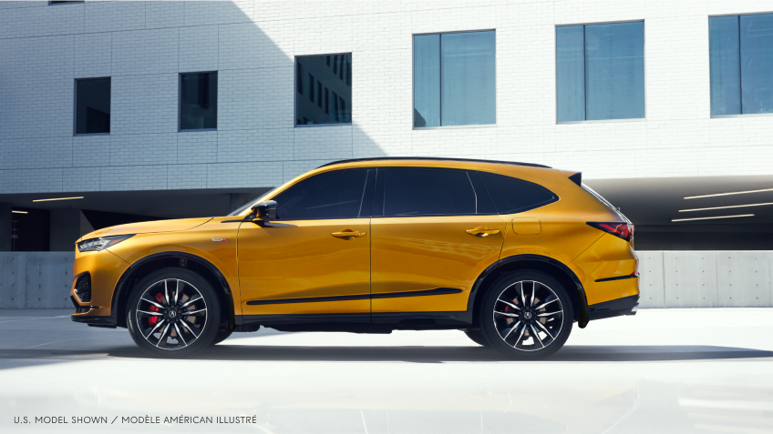 Wide side view of gold MDX Type S parked in a parking lot outside a modern, minimalistic white building.